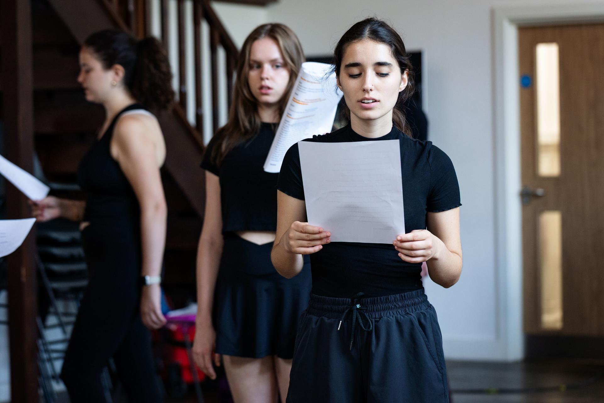 Performers dressed in black read from scripts during a rehearsal in a hallway